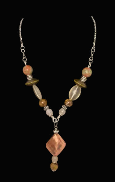 Made using semi precious stones and a copper distressed main bead that is hollow and approx. 1 1/4". A flying saucer shaped bead, a marbleized semi-precious 10mm bead "rounds" out the look; on a Black velvet necklace bust on a white background.