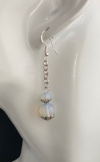 A single dangling earring displayed on a white ceramic mannequin head against a black background. The earring has a silver-toned chain and hook with a small iridescent bead that shifts between pale blue, white, and hints of pink.