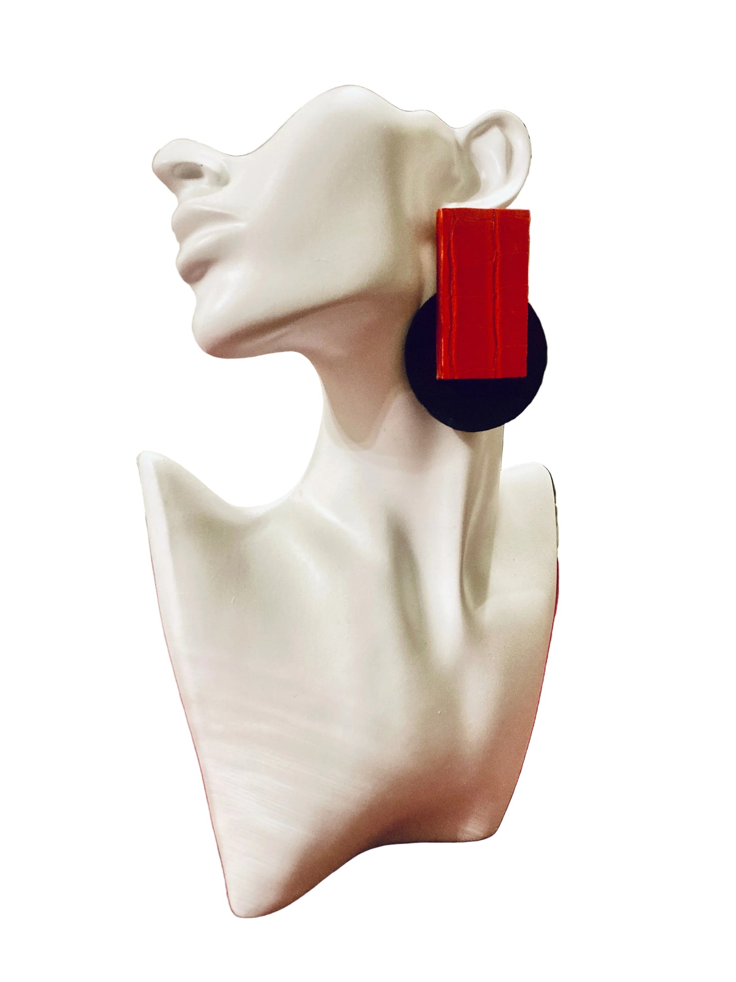 A pair of large geometric statement earrings displayed on a white ceramic female bust mannequin. The earrings feature a bold rectangular red top and a wide black circular base, set against a white background.