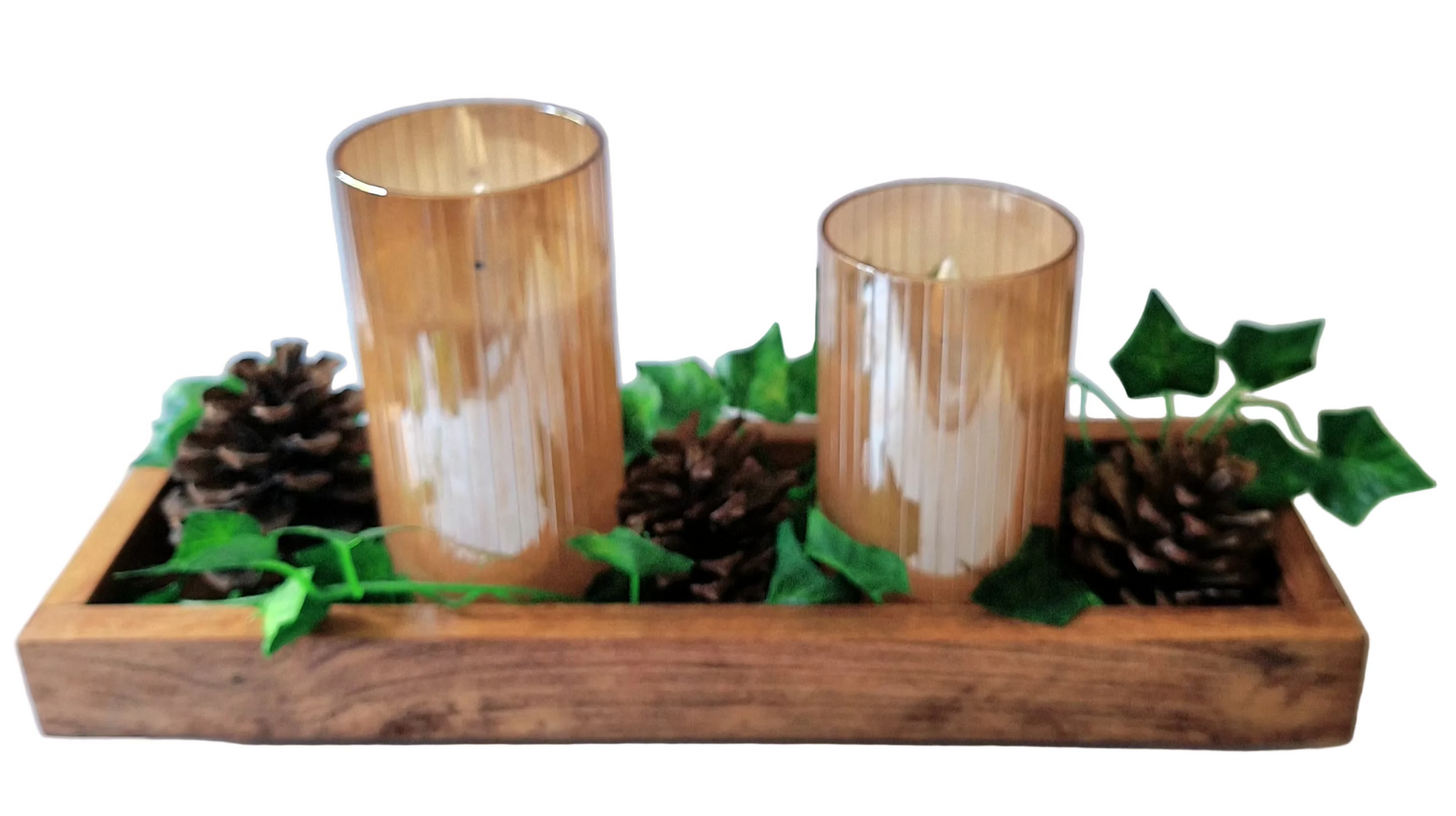A rustic holiday centerpiece wooden tray with two amber glass candle holders, with white LED candles, with green ivy and pinecones against a plain beige wall.