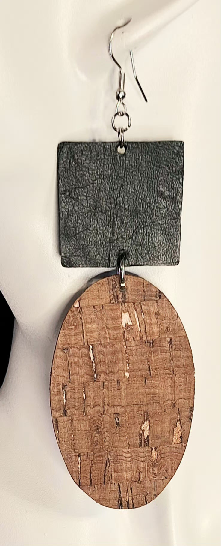 A pair of handmade dangle earrings featuring a textured black square top and a natural cork oval bottom, suspended from silver fishhook wires on a white background.