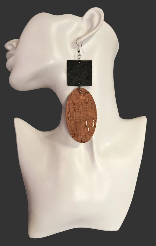 A pair of statement earrings displayed on a white mannequin bust. One earring features a square black stone top with a silver hook, and the other features a large oval cork-textured faux leather drop.
